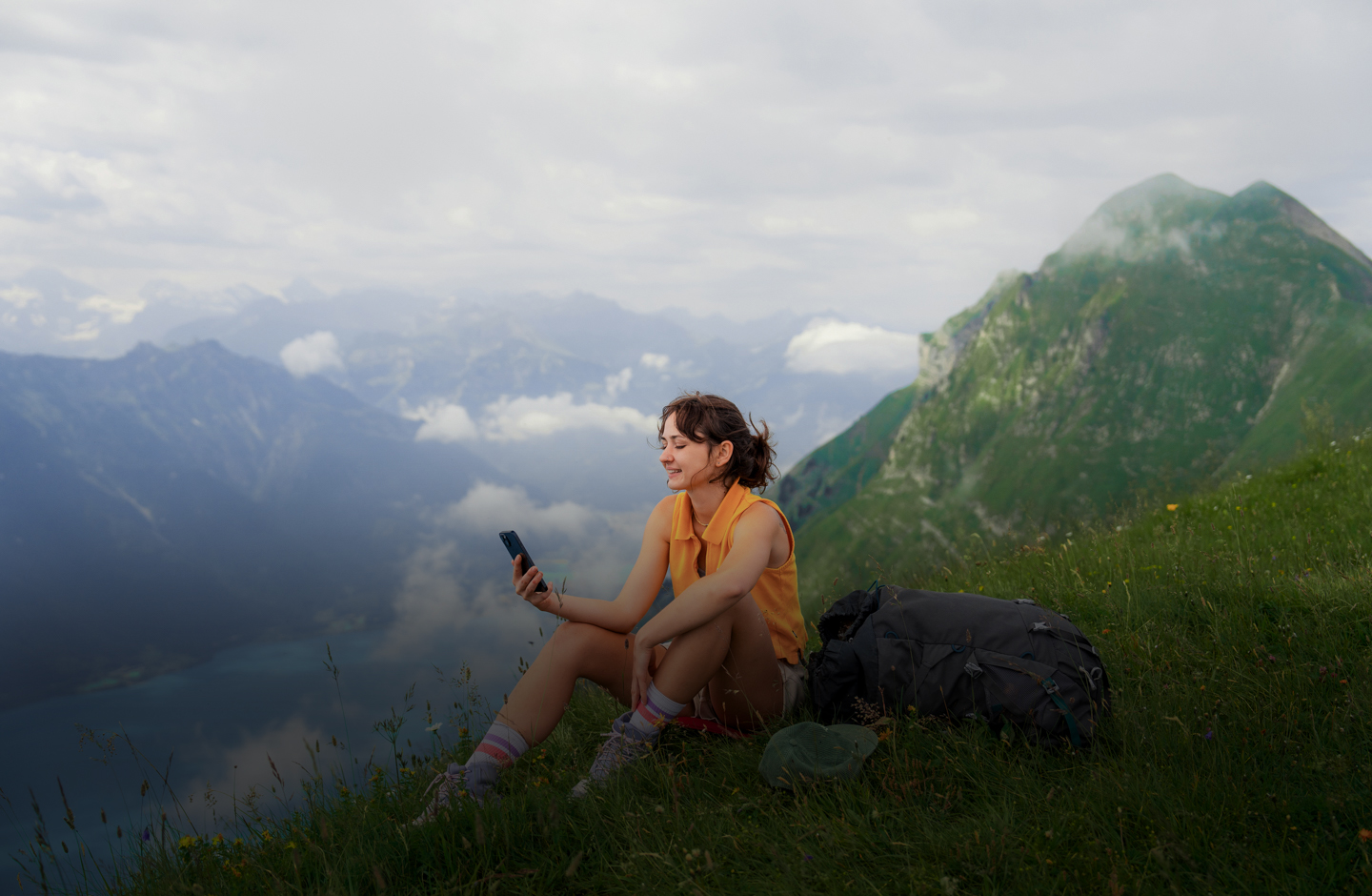 Eine junge Frau sitzt auf einer Wiese am Gipfel eines Berges und blickt auf ihr Smartphone.